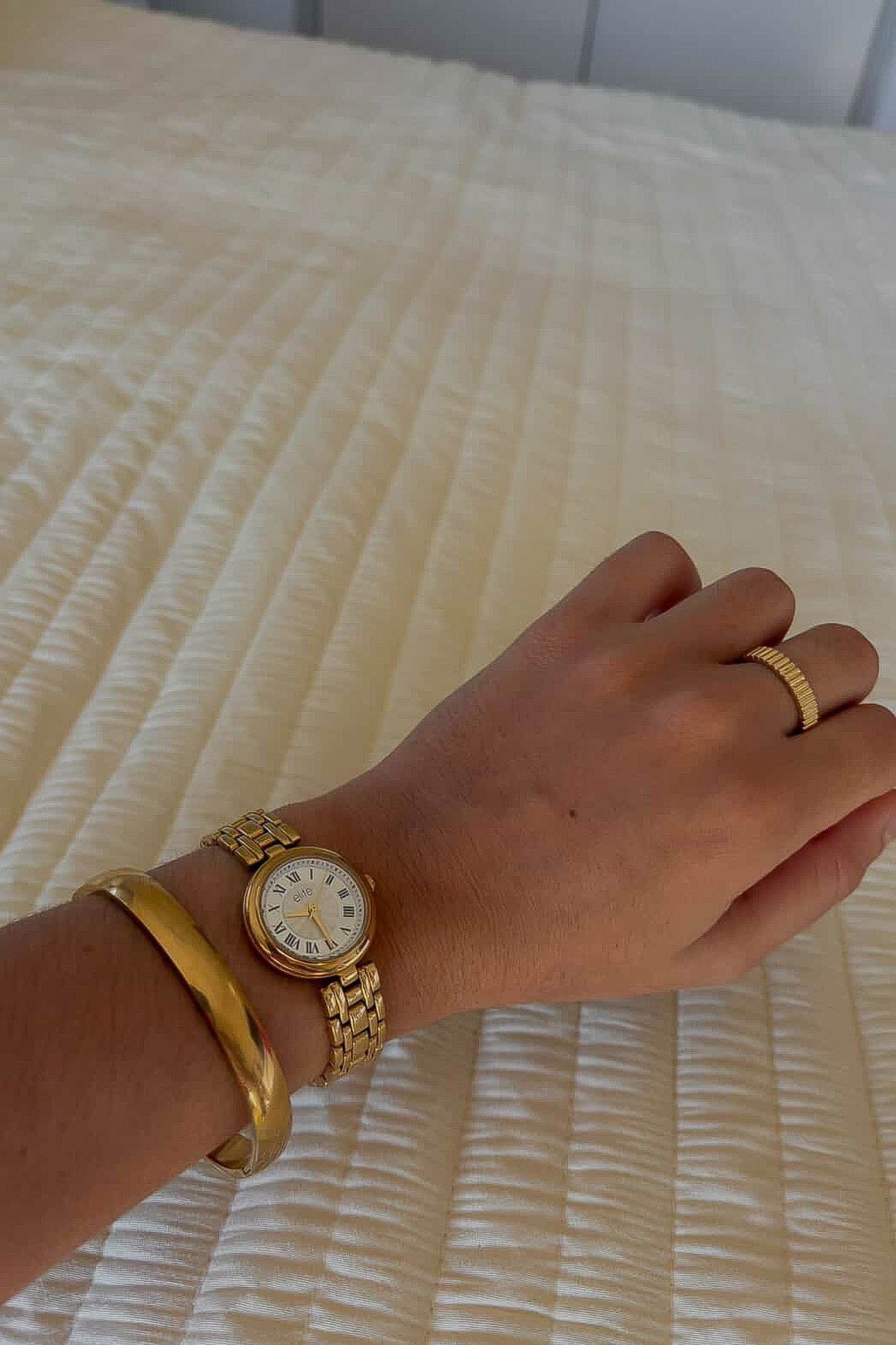 Hand wearing a gold watch and bracelet on a textured surface