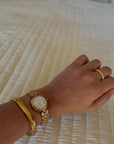 Hand wearing a gold watch and bracelet on a textured surface