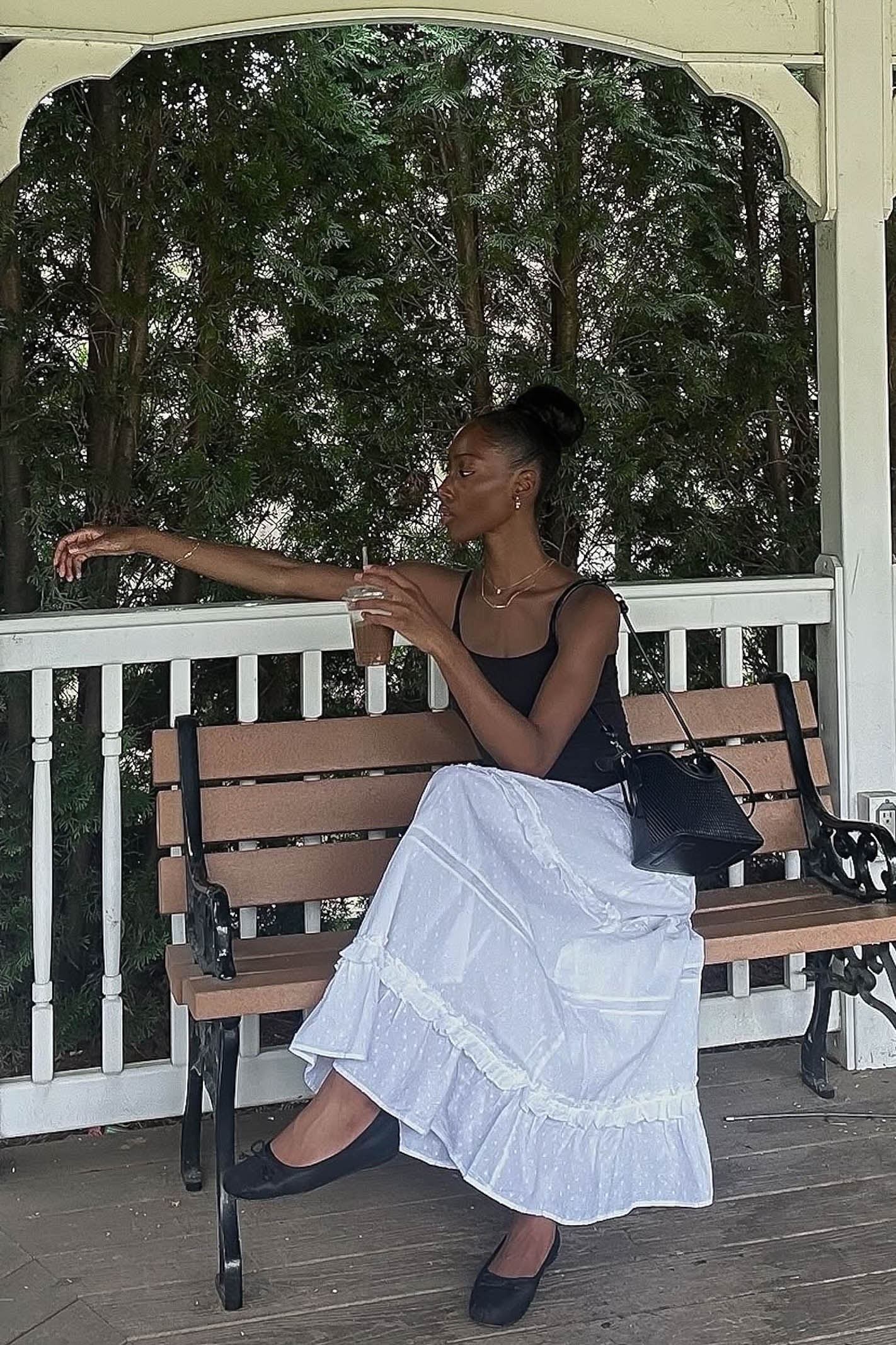 Woman sitting on a bench holding a drink, with trees and a white railing in the background