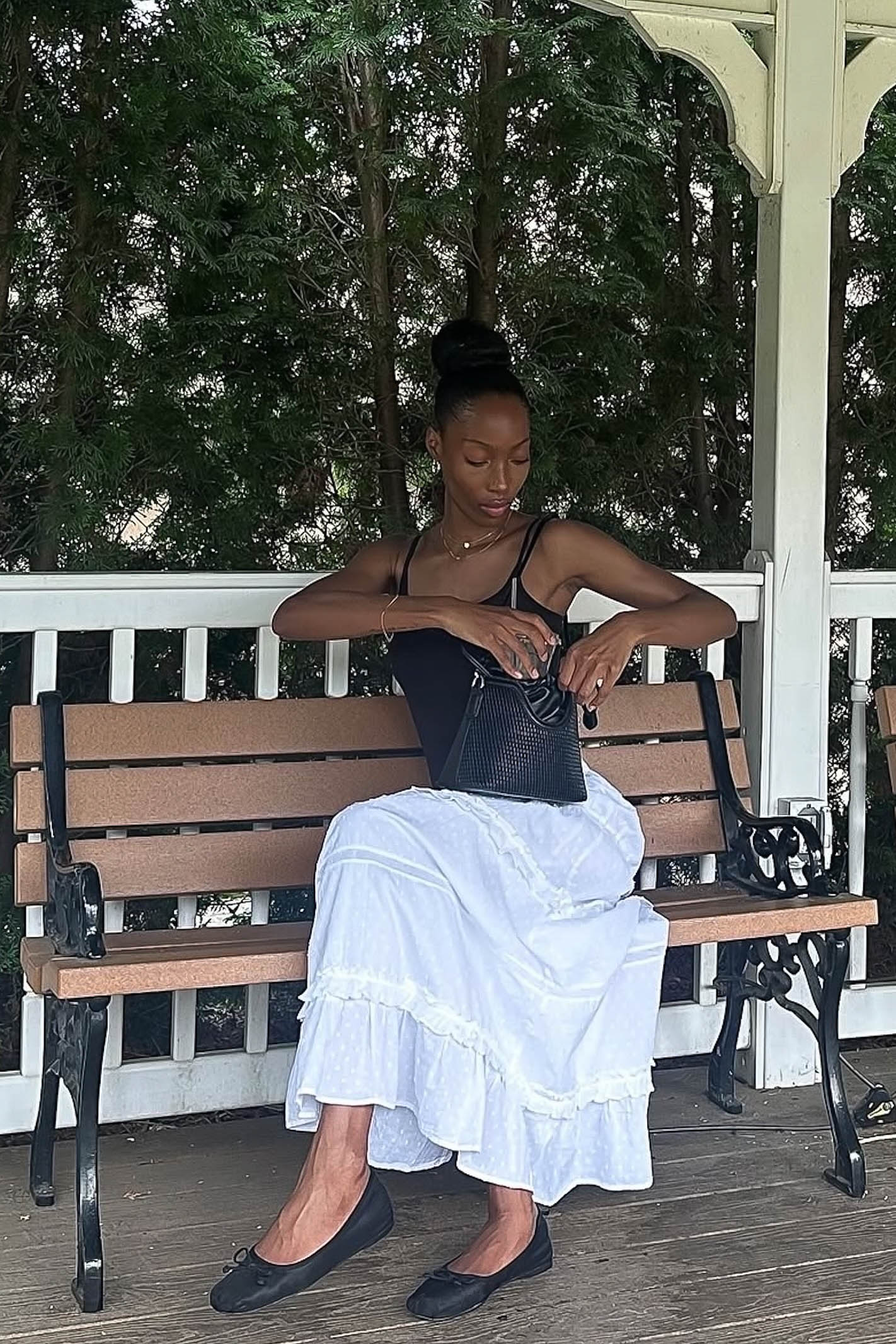 Woman sitting on a bench outdoors, wearing a black top and white skirt.