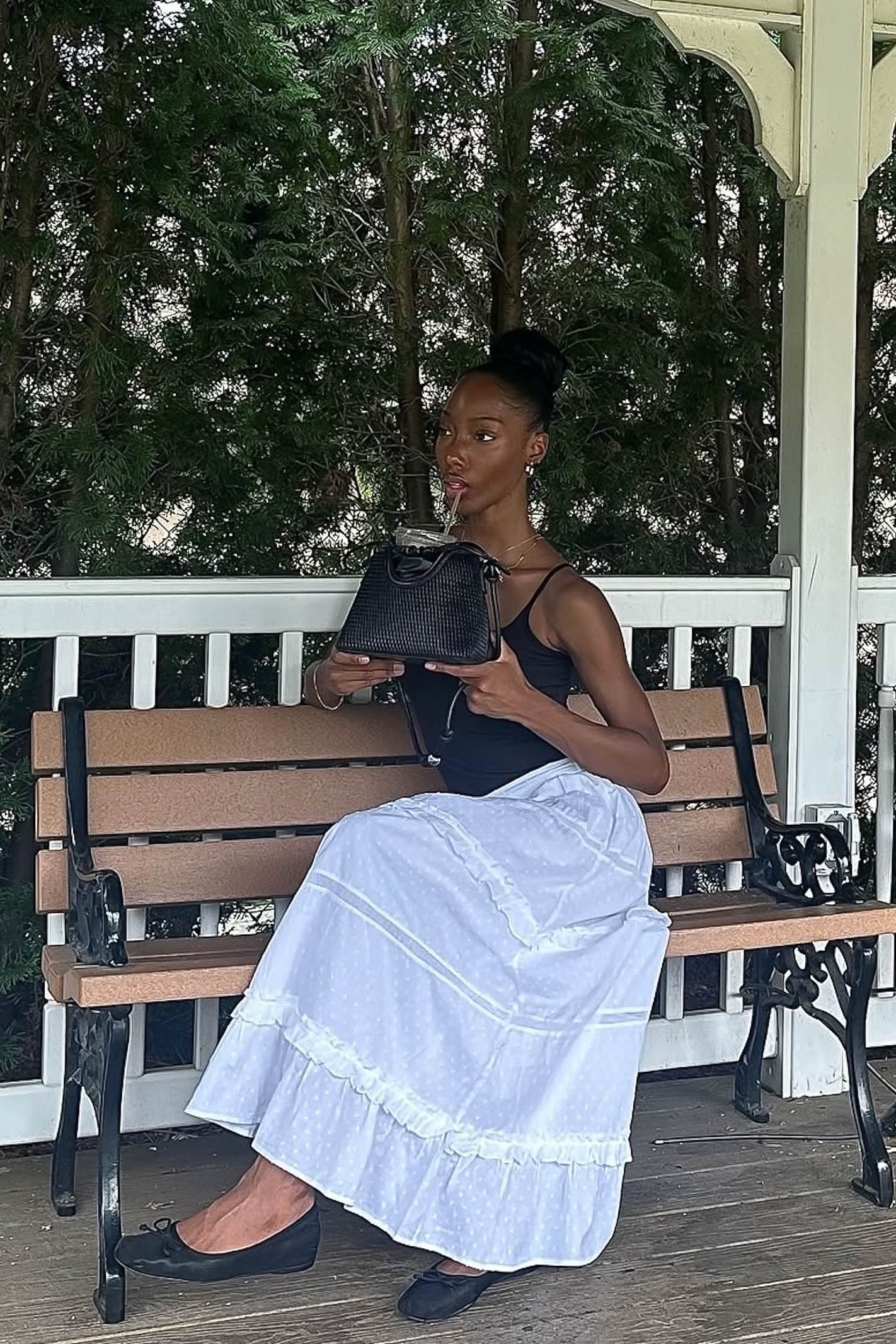 Woman sitting on a bench holding a black handbag, wearing a black top and white skirt.