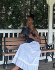 Woman sitting on a bench holding a black handbag, wearing a black top and white skirt.
